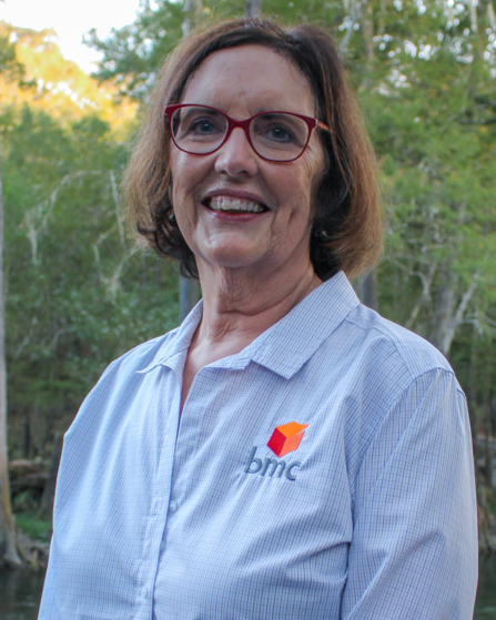 Dr. Riordan standing outside near trees smiling, wearing a blue company button down shirt and red glasses