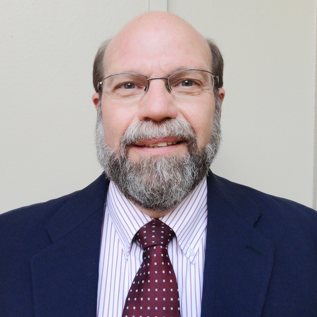 Dr. Coleman smiling with a white beard while wearing glasses, a pink button down and black blazer